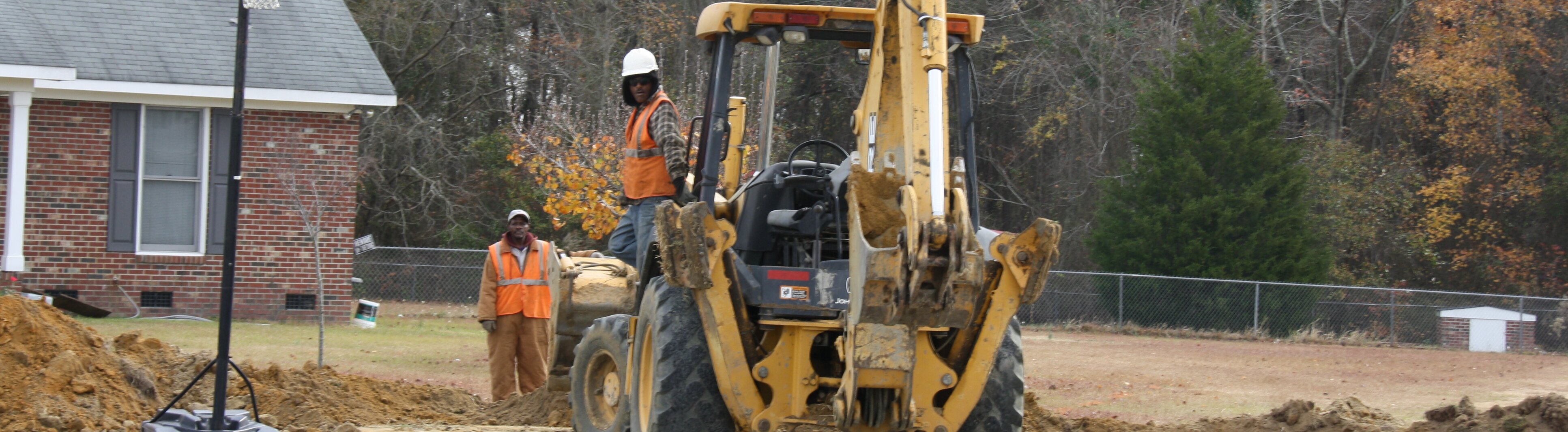 Two construction workers operate heavy equipment near a brick house, digging with a backhoe in a yard.